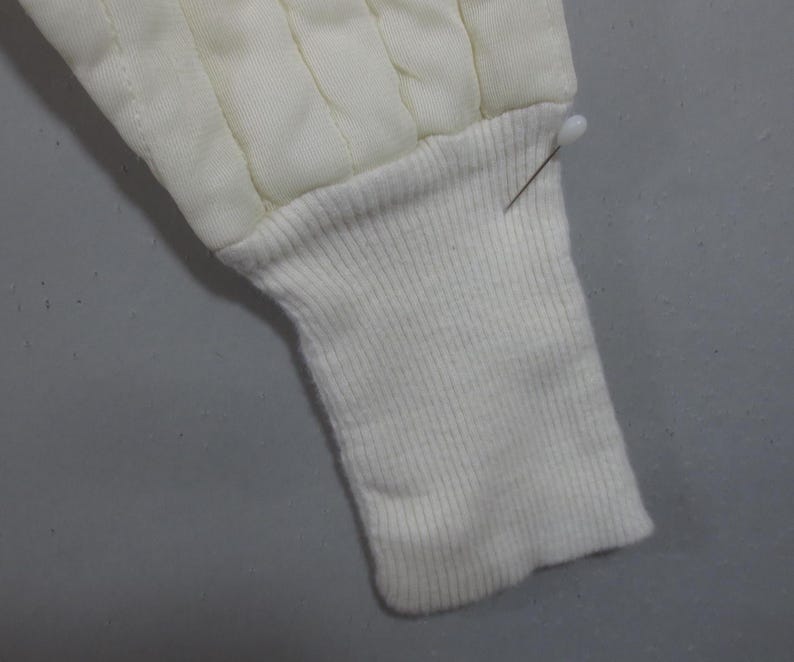 May include: Close-up of a white quilted jacket sleeve with a ribbed knit cuff. The cuff is folded back and secured with a pin.