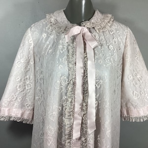 May include: A vintage, light pink lace robe with short sleeves. The robe features a delicate floral pattern, a ruffled collar, and a satin ribbon tie. The sleeves and front edges are trimmed with lace.