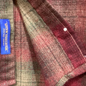 Puede incluir: Primer plano de una tela a cuadros en tonos rojos, verdes y marrones. Una etiqueta azul con texto blanco que dice "HIGH GRADE WESTERN WEAR" est&aacute; cosida a la tela. Se ve un alfiler blanco.