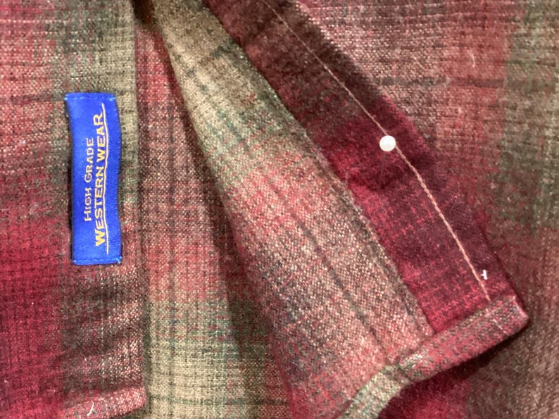 Puede incluir: Primer plano de una tela a cuadros en tonos rojos, verdes y marrones. Una etiqueta azul con texto blanco que dice "HIGH GRADE WESTERN WEAR" est&aacute; cosida a la tela. Se ve un alfiler blanco.