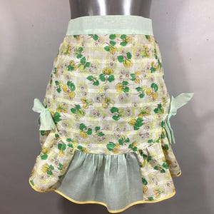 May include: A vintage green and white floral print apron with a ruffled hem and yellow trim. The apron has two green bows on the sides.