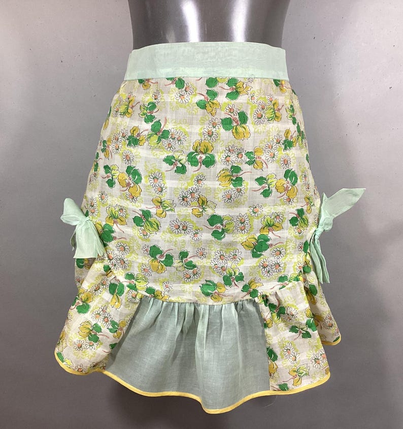 May include: A vintage green and white floral print apron with a ruffled hem and yellow trim. The apron has two green bows on the sides.