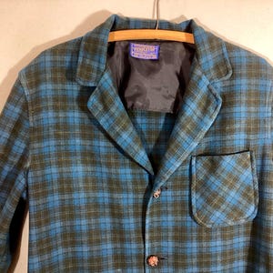 May include: A blue and green plaid blazer with a single pocket and two buttons. The blazer is hanging on a wooden hanger. The label inside the blazer reads "Pendleton".