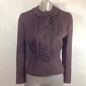 May include: A dark brown, vintage jacket with a rounded collar and four matching buttons down the front. The jacket has long sleeves and small pockets on each side. The jacket is displayed on a mannequin.