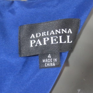 K&ouml;nnte beinhalten: Ein blaues Kleider-Etikett mit dem Text "Adrianna Papell" und "Made in China" in wei&szlig;er Schrift. Das Etikett tr&auml;gt au&szlig;erdem die Nummer "4".