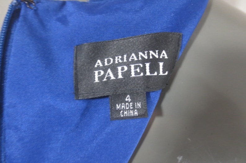 K&ouml;nnte beinhalten: Ein blaues Kleider-Etikett mit dem Text "Adrianna Papell" und "Made in China" in wei&szlig;er Schrift. Das Etikett tr&auml;gt au&szlig;erdem die Nummer "4".
