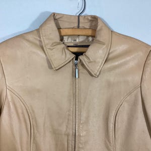 May include: A tan leather jacket with a collared neckline and a full-length zipper. The jacket has a classic design with seams that run down the front and sleeves. The jacket is hanging on a wooden hanger.
