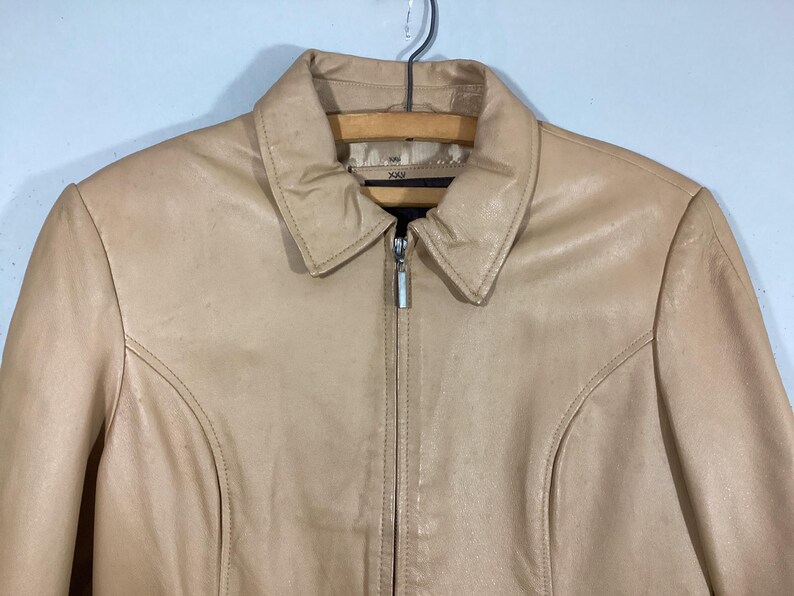 May include: A tan leather jacket with a collared neckline and a full-length zipper. The jacket has a classic design with seams that run down the front and sleeves. The jacket is hanging on a wooden hanger.