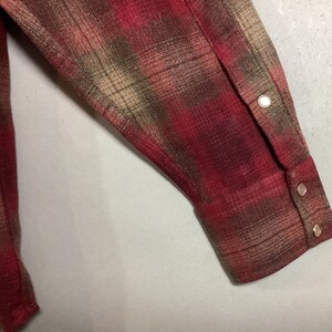 Puede incluir: Primer plano de una manga de camisa a cuadros roja, marr&oacute;n y beige. La manga tiene un pu&ntilde;o abotonado con dos cierres de presi&oacute;n plateados. La tela tiene un aspecto texturizado.