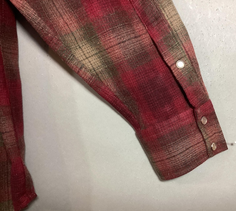 Puede incluir: Primer plano de una manga de camisa a cuadros roja, marr&oacute;n y beige. La manga tiene un pu&ntilde;o abotonado con dos cierres de presi&oacute;n plateados. La tela tiene un aspecto texturizado.