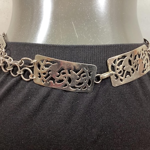 May include: A silver chain belt with two large, ornate, rectangular, cutout metal plates. The plates have a floral design.