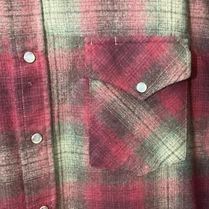 Puede incluir: Primer plano de una camisa a cuadros rojos y verdes con dos bolsillos en el pecho y cierres de bot&oacute;n a presi&oacute;n plateados. La camisa tiene un estilo occidental cl&aacute;sico con una parte delantera abotonada y una tela suave y texturizada.