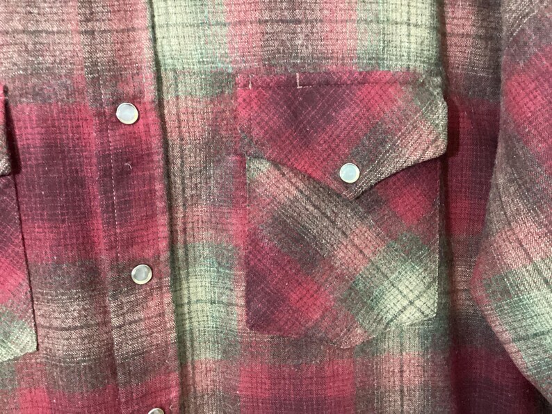 Puede incluir: Primer plano de una camisa a cuadros rojos y verdes con dos bolsillos en el pecho y cierres de bot&oacute;n a presi&oacute;n plateados. La camisa tiene un estilo occidental cl&aacute;sico con una parte delantera abotonada y una tela suave y texturizada.