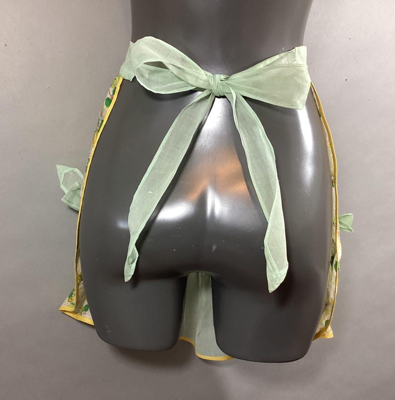 May include: A yellow and green floral print apron with a light green sheer ribbon tie. The apron has a gathered skirt and two side pockets.