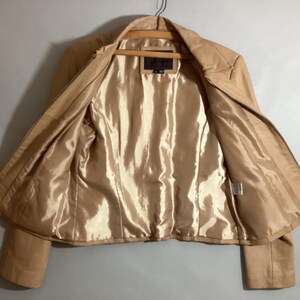 May include: A tan leather jacket with a full zipper closure, hanging on a wooden hanger. The interior lining is a shiny, golden-tan color. The jacket is open, displaying the interior lining and the zipper.