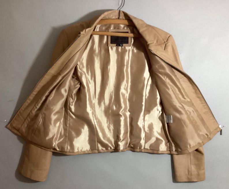 May include: A tan leather jacket with a full zipper closure, hanging on a wooden hanger. The interior lining is a shiny, golden-tan color. The jacket is open, displaying the interior lining and the zipper.