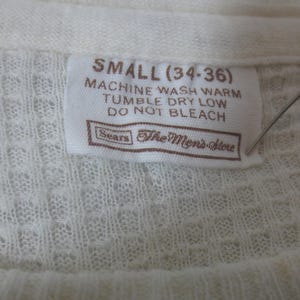 May include: A white tag with brown text that reads "SMALL (34-36)" and "MACHINE WASH WARM TUMBLE DRY LOW DO NOT BLEACH" with the Sears logo and "The Men's Store" below the text.