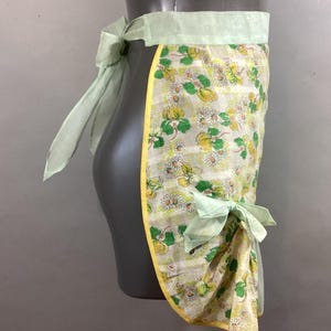 May include: A vintage green and white floral print apron with yellow trim and light green ties. The apron is tied at the waist and has a bow on the side.