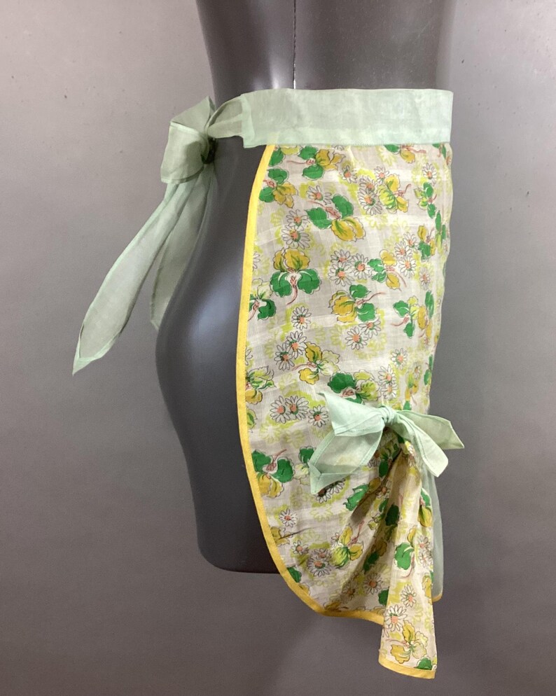 May include: A vintage green and white floral print apron with yellow trim and light green ties. The apron is tied at the waist and has a bow on the side.