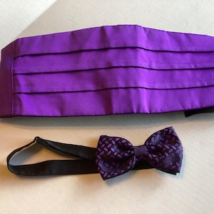 May include: A vibrant purple cummerbund featuring horizontal pleats paired with a matching black bow tie that has a repeating purple oval pattern. The bow tie includes an adjustable black strap for securing around the neck, completing the set.