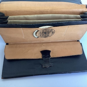 May include: A black leather briefcase with a brown leather interior. The briefcase has a flap closure with a metal clasp and a circular metal design on the front.