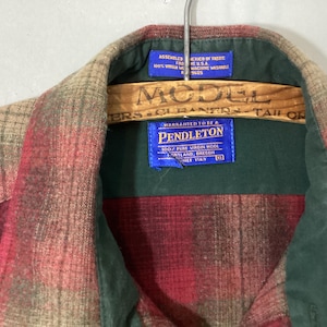 Puede incluir: Una camisa a cuadros Pendleton en tonos rojos, beige y verdes, colgada de una percha de madera. La camisa tiene un cuello verde y una etiqueta azul con el logotipo y el texto de Pendleton.