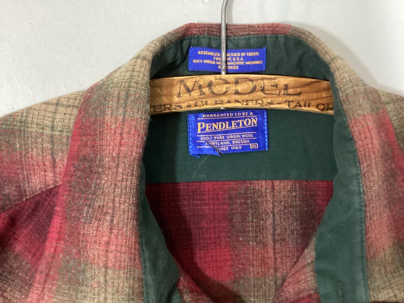 Puede incluir: Una camisa a cuadros Pendleton en tonos rojos, beige y verdes, colgada de una percha de madera. La camisa tiene un cuello verde y una etiqueta azul con el logotipo y el texto de Pendleton.