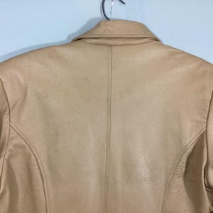 May include: A tan leather jacket, viewed from the back, hangs on a metal hanger. The jacket features a classic collar, shoulder seams, and a vertical seam down the center of the back. The leather has a slightly worn appearance.