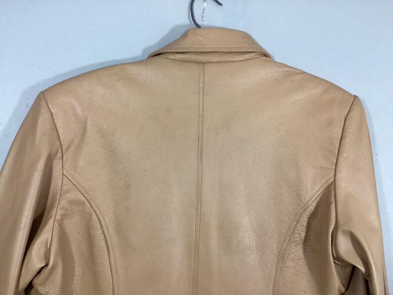 May include: A tan leather jacket, viewed from the back, hangs on a metal hanger. The jacket features a classic collar, shoulder seams, and a vertical seam down the center of the back. The leather has a slightly worn appearance.