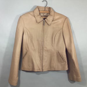 May include: A tan leather jacket with a collared neckline and a full-length zipper. The jacket has long sleeves, two front pockets, and is hanging on a metal hanger. The jacket is a neutral color and appears to be in good condition.