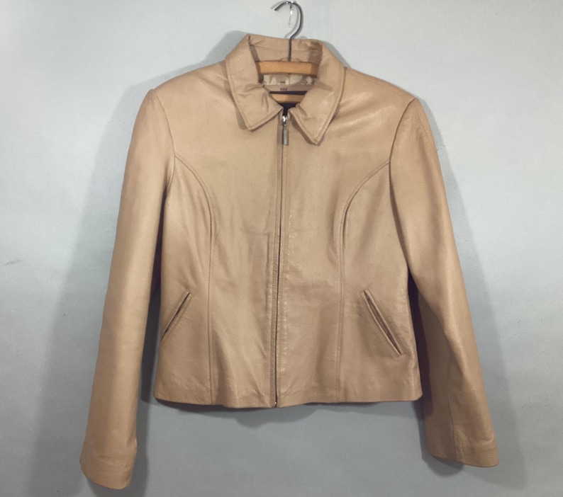 May include: A tan leather jacket with a collared neckline and a full-length zipper. The jacket has long sleeves, two front pockets, and is hanging on a metal hanger. The jacket is a neutral color and appears to be in good condition.