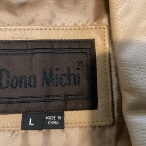 May include: Close-up of a beige leather jacket with a label that reads "Dona Michi" in dark brown lettering. The label also indicates the size "L" and "MADE IN CHINA". The jacket has a quilted lining.