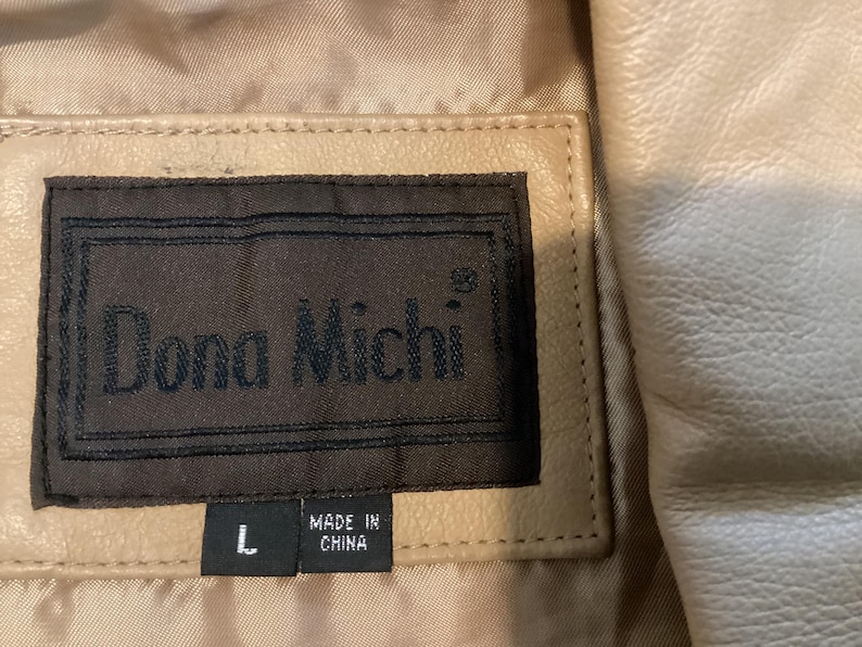 May include: Close-up of a beige leather jacket with a label that reads "Dona Michi" in dark brown lettering. The label also indicates the size "L" and "MADE IN CHINA". The jacket has a quilted lining.