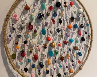 Può includere: Un acchiappasogni rotondo con un centro a rete metallica, adornato con una varietà di perline colorate in tonalità di rosso, blu, verde e oro. L'anello esterno è avvolto in spago naturale e c'è un anello di spago nella parte superiore per appenderlo.