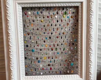 Può includere: Espositore di gioielli con cornice bianca ornata con un retro in rete metallica. La rete contiene numerose perline colorate di varie forme e dimensioni, creando un pezzo vibrante e decorativo. La cornice ha intagli dettagliati.