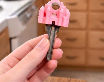 May include: A metal key featuring a light pink, three-dimensional plastic topper shaped like a castle with multiple towers and a pointed roof. The castle topper is attached to the bow of the key, which has a standard silver-colored metal blade.