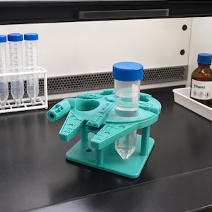 Könnte beinhalten: Ein teal-farbener, 3D-gedruckter Reagenzglasständer in Form des Millennium Falcon mit einem transparenten Reagenzglas, das Flüssigkeit enthält. Weitere Reagenzgläser und eine Flasche Ethanol sind im Hintergrund zu sehen.