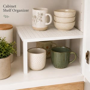 Puede incluir: Un organizador de estante de armario de metal blanco con una superficie superior perforada. El texto 'Cabinet Shelf Organizer' está impreso en la esquina superior izquierda. Sostiene una taza floral y tres cuencos apilados, con dos tazas debajo.