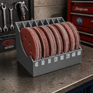 May include: A gray plastic storage organizer holding multiple circular red sandpaper discs. The organizer features vertical slots and white printed numbers 40, 60, 80, 120, 180, 220, 400, and 600 along the front edge to categorize the grit levels of the abrasive discs.