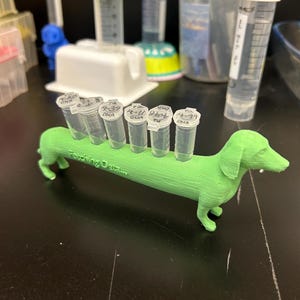 May include: A green 3D-printed dachshund test tube holder, holding six clear tubes labelled "DNA". The holder has the text "Fetching Data" printed on its side. Other lab equipment is visible in the background.