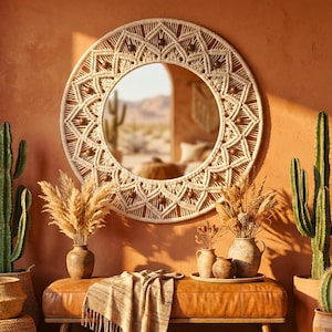 Può includere: Uno specchio rotondo in macramè color crema con un design geometrico e perline di legno. È appeso a una parete color terracotta, sopra una panca in pelle con vasi ed erba della pampa. I cactus sono ai lati.