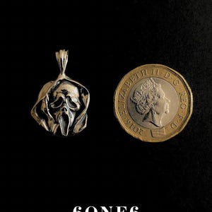 May include: A silver pendant in the shape of a Scream mask, next to a British one-pound coin. The pendant has a detailed, textured design. The coin shows a profile of Queen Elizabeth II. The text "6ONE6 JEWELLERY" is at the bottom.