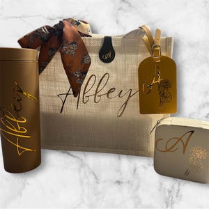 May include: A collection of personalized travel accessories, including a brown tumbler, a tote bag with the name "Abbey", a luggage tag, and a small cosmetic bag. The items are in shades of brown and beige, with gold accents.