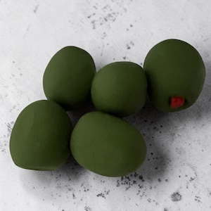 May include: Five green olives with a matte finish are arranged on a light gray surface. One olive has a small red pimiento visible. The olives are a uniform size and shape, suggesting they are a food item.