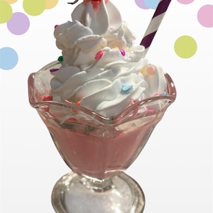 Puede incluir: Un batido rosa en una copa de helado de cristal transparente, cubierto con crema batida, chispas y una cereza roja. Una pajita a rayas moradas y blancas está insertada en el batido. El fondo presenta lunares de colores.