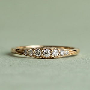 Peut inclure: Une bague en or avec sept petites pierres précieuses rondes et claires disposées en ligne. La bague présente un motif délicat et détaillé sur les bords. L'arrière-plan est vert clair.