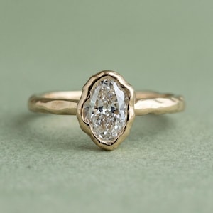 May include: A gold ring with a large, oval-shaped diamond. The diamond is set in a bezel setting with a textured, organic design. The band of the ring is also textured, giving it a unique, handcrafted look.