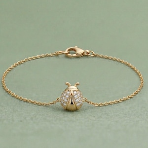 Peut inclure: Un bracelet doré avec un charm en forme de coccinelle. La coccinelle est ornée de petits pierres précieuses transparentes. Le bracelet a une chaîne délicate et une fermeture à mousqueton. Le fond est vert.