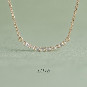 May include: A gold chain necklace featuring a curved bar pendant set with alternating baguette and round cut clear stones. The necklace is displayed above the word LOVE printed in black text. The design is symmetrical and delicate in appearance.