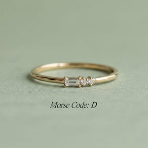 Op de afbeelding: Een dunne gouden ring met een horizontale baguette-geslepen diamant gevolgd door twee kleine briljant-geslepen diamanten. De tekst Morse Code: D staat onder de ring. Het ontwerp bevat drie stenen die de letter D in morsecode weergeven.
